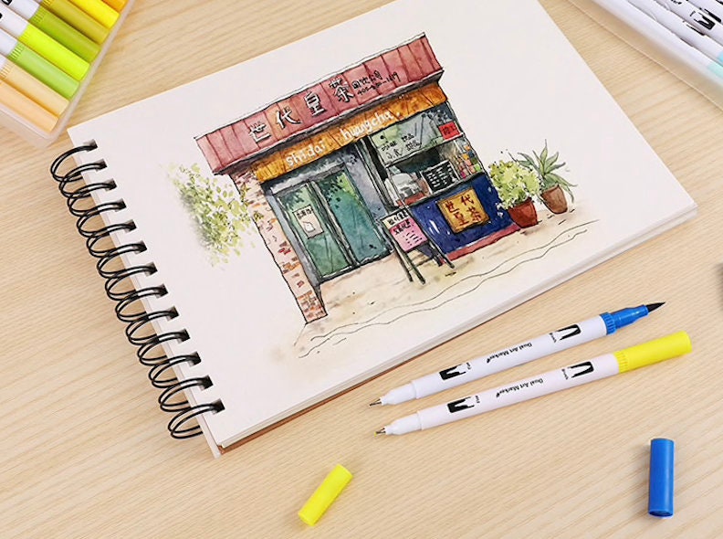 36 Colors Dual Tip Brush Markers Set for Art with detailed drawing of a storefront on open sketchbook, yellow and blue markers on wood table