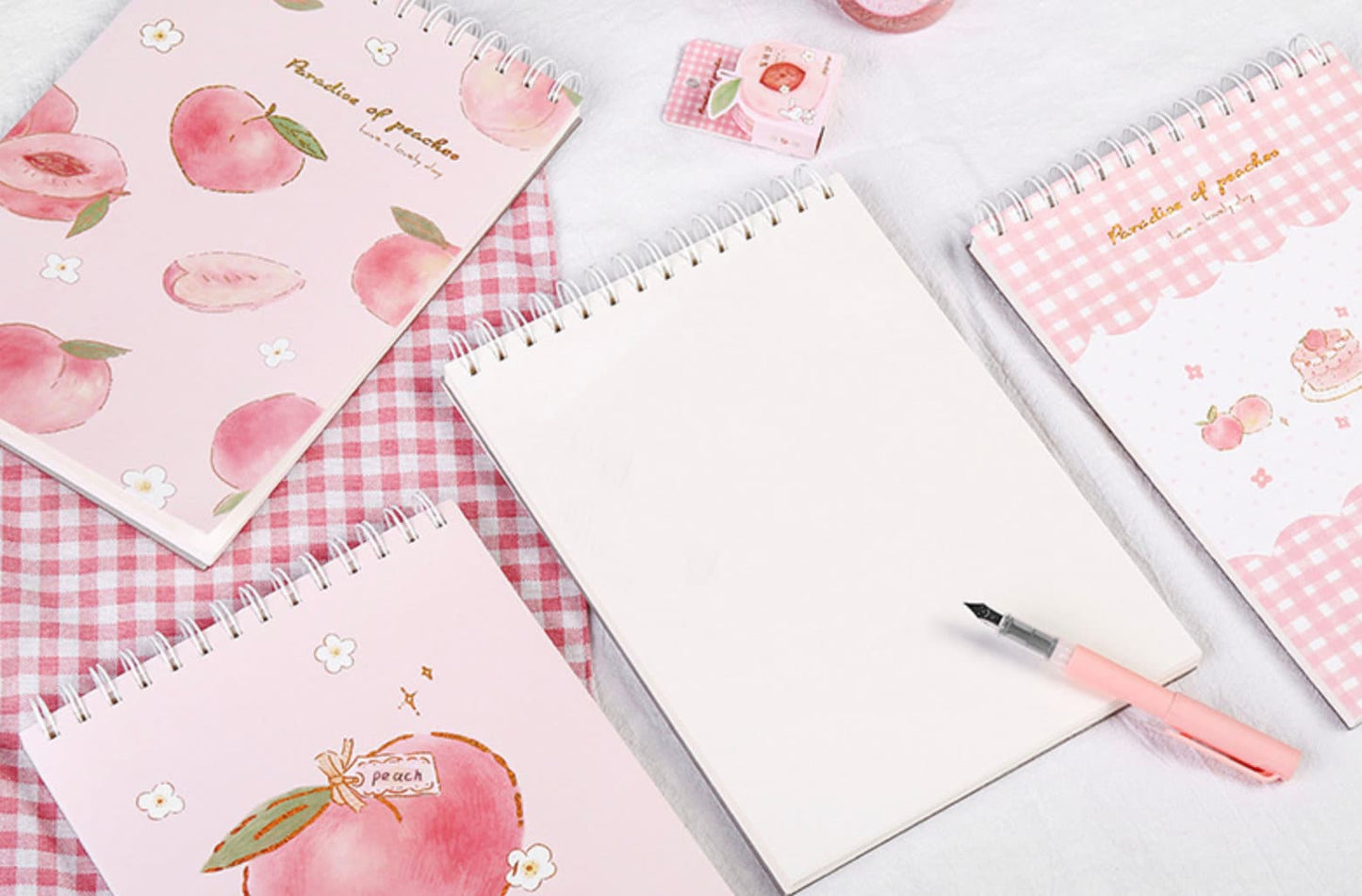 A4 Pink Peach Coil Sketch Book - Cute Planner with adorable peach illustrations and pink covers. Perfect for journaling and drawing. Lies on a pink checkered cloth with a peach-themed rubber, tape, and a pink pen beside it. Ideal for kawaii stationary lovers.