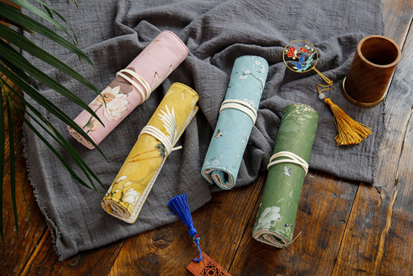 Floral Roll-Up Pencil Case - Artist Brush Holder in pastel pink, mustard yellow, light blue, and forest green with elegant floral designs lying on a textured grey cloth on a rustic wooden table with decorative elements