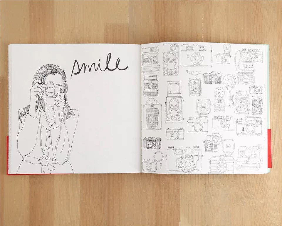 Open Traveling Paris Coloring Book for Kids and Adults showing pages with "Smile" and various camera illustrations, on a wooden table.