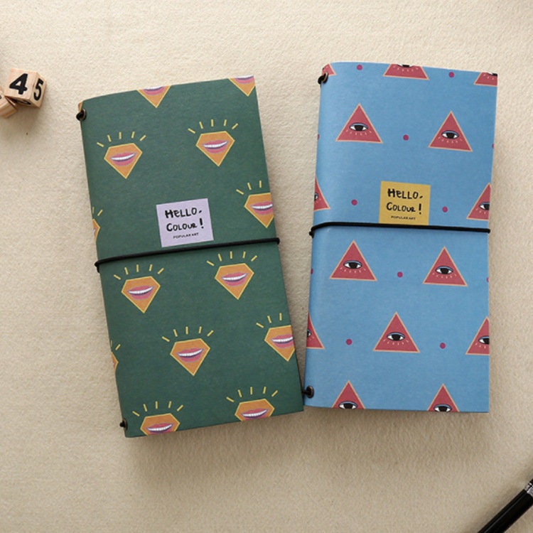 Spring Flower Kraft Journals - Recycled Paper Set featuring colorful and quirky designs. Two notebooks are shown: one with a green cover and smiling mouth pattern, and the other with a blue cover and eye-triangle motif. Eco-friendly and stylish stationery for daily use.