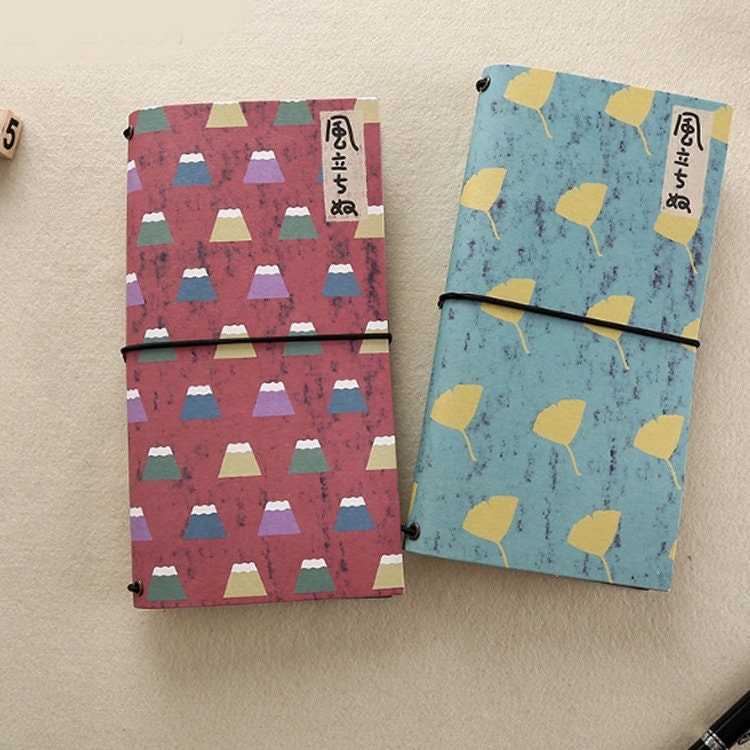 Spring Flower Kraft Journals - Recycled Paper Set featuring two journals, one with a pink background and pastel mountain pattern, the other with a blue background and yellow leaf design, secured with black elastic bands.