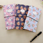 Spring Flower Kraft Journals - Recycled Paper Set featuring three journals with floral designs in pink, purple, and blue covers and a black pen beside them.