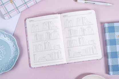 Hard Cover A5 Minimalist Grid Pocket Planner open on a pink desk, surrounded by a blue patterned cover book, a white pen, and a light blue decorative plate.