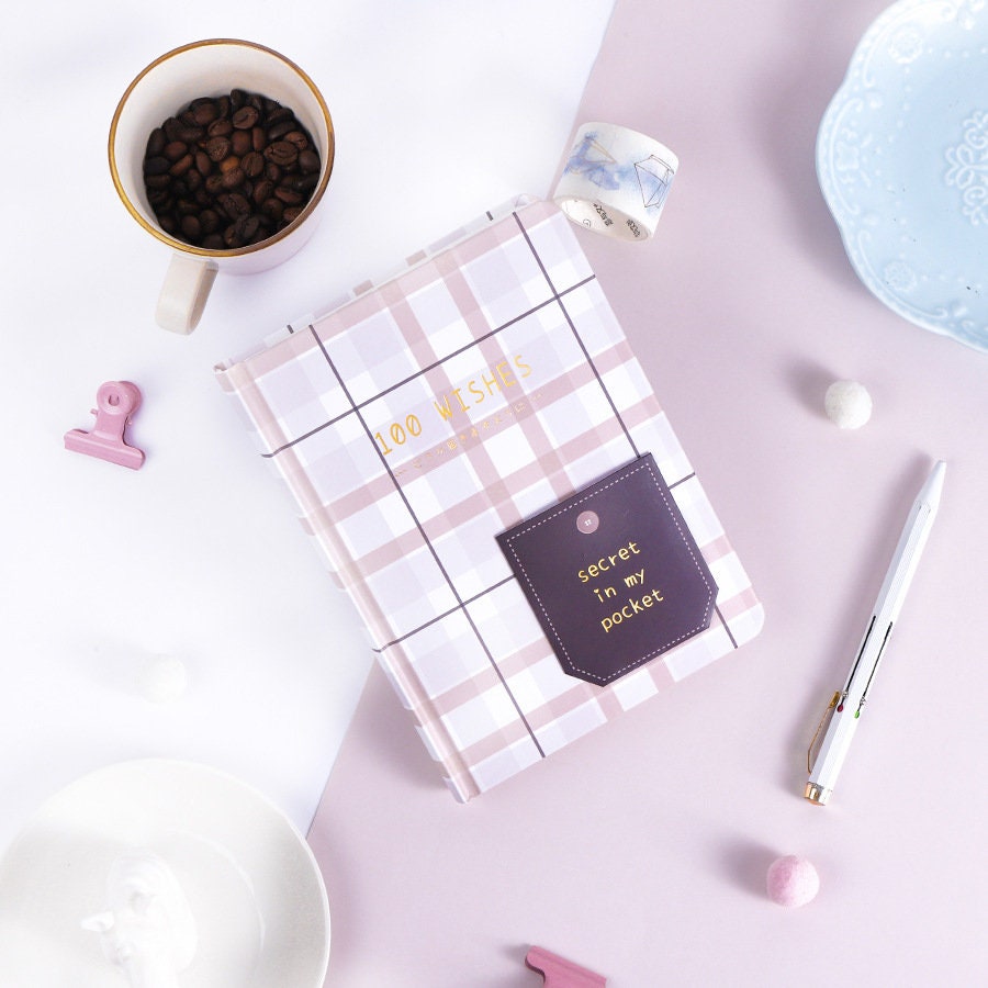 Hard Cover A5 Minimalist Grid Pocket Planner with "100 Wishes" and "Secret in my pocket" text, surrounded by stationery items, coffee cup filled with beans, and decorative items on a pastel background.