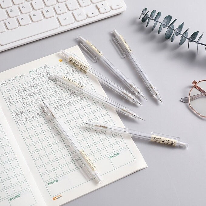 0.5mm transparent mechanical pencils for office and student stationery arranged next to an open notebook with handwritten text, surrounded by office items including a keyboard, glasses, and plant decor.