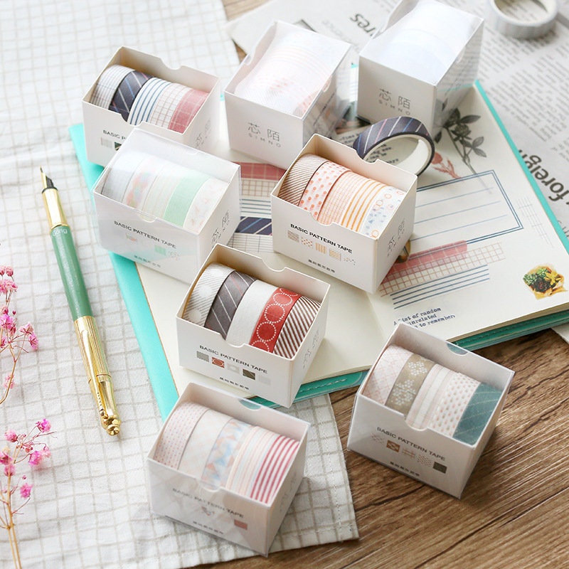 Cute Colorful Washi Tape Set in 5 Rolls, 5 Meters each, displayed in boxes with various pastel and patterned designs on a desk with stationary and a green pen.