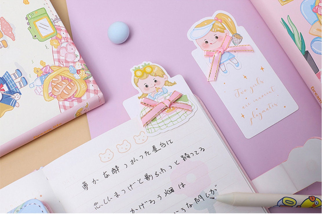 Pink leather journal with magnetic buckle, bookmark with cute cartoon girl illustration, pastel-colored stationery, kawaii style, perfect for note-taking and journaling.