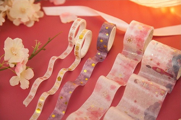 Japanese Cherry Blossom Washi Tape Set - 6 Rolls, featuring pastel pink, purple, and white tapes with delicate floral and gold foil accents, displayed on a pink background with cherry blossom flowers. Perfect for decorating planners, journals, and crafts.