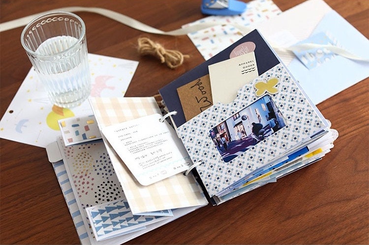 Aesthetic scrapbook paper and decorative journaling supplies displayed on a wooden table next to a glass of water, featuring a scrapbook with various patterned papers, photos, and ephemera.