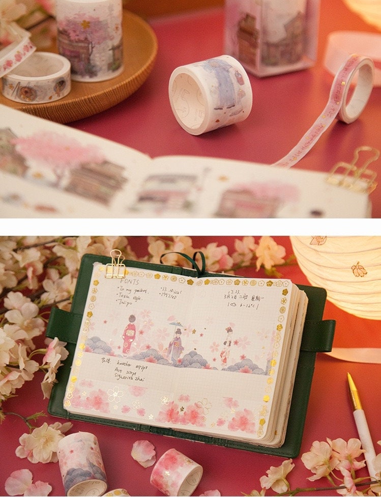 Japanese Cherry Blossom Washi Tape Set - 6 Rolls displayed on a pink background, with open journal pages decorated with the washi tape. The tapes feature cherry blossom designs, traditional Japanese illustrations, and pastel colors. Perfect for bullet journaling, scrapbooking, and adding a kawaii touch to planners and crafts.