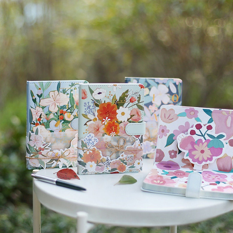 Pink Floral Garden Planner - 196 Pages Leather Notebook with colorful flower designs on the cover, displayed on a white table in a garden setting, alongside a pen and decorative leaf stickers. Perfect for organizing and journaling.