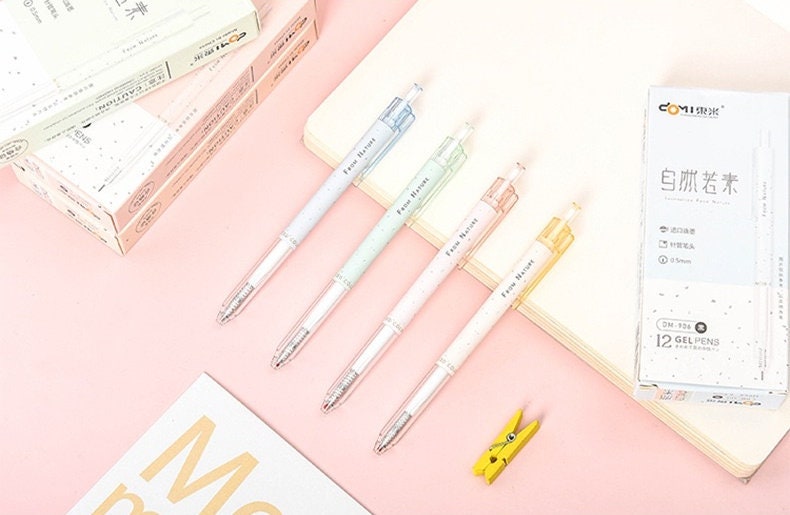 Minimalist 0.5mm black ink gel pens with different colored grips for office and student use, displayed on a pink desk with notebooks and packaging boxes in the background.