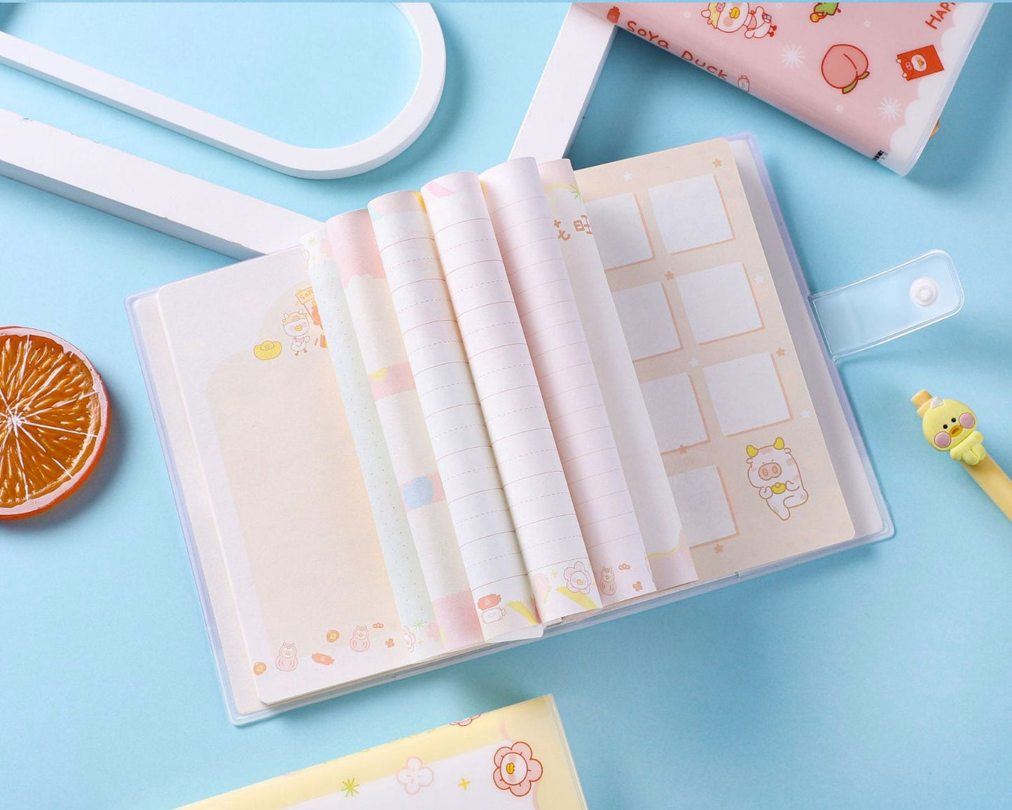 Soya Duck Leather Notebook - A5, 224 Pages with kawaii illustrations on pastel-colored pages, shown partially open against a blue background with a cute turtle pen and a sliced orange decoration.