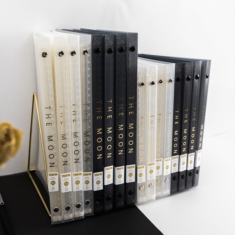 B5 Stars and Moons PVC Cover Planner in black and clear, displayed on a gold wire stand, featuring the text "The Moon" on the spine and gold embellishments.