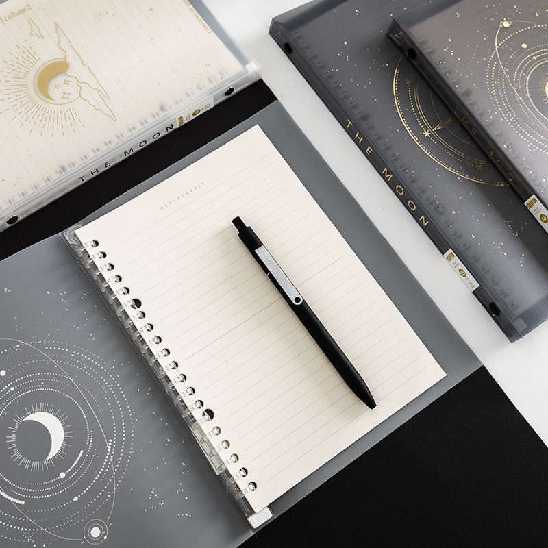 B5 Stars and Moons PVC Cover Planner with celestial design, open with black pen on top, featuring spiral binding and lined pages.