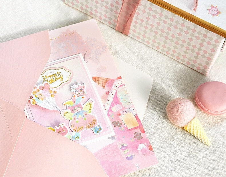 3D Pink Cake Pop Up Card - Happy Birthday, kawaii style with cute illustrations, in a pink envelope, surrounded by pastel-colored macarons and decorative items.