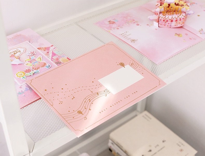 3D Pink Cake Pop Up Card - Happy Birthday on a white shelf, surrounded by other cute and colorful kawaii stationery items. The card is light pink with gold detailing and musical notes.