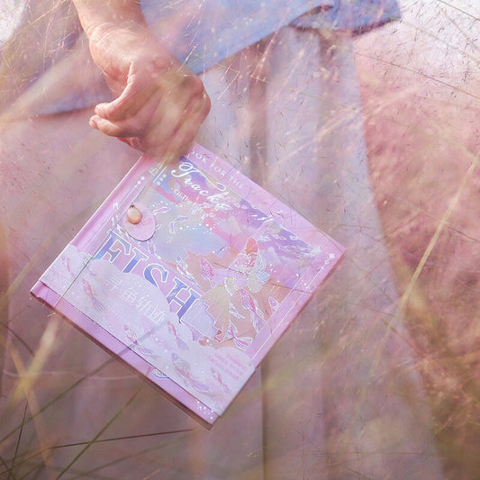 Fish Hear Sea Hard Cover Journal with Bookmark in pastel colors of pink and purple with whimsical illustrations, held by a hand amidst dreamy soft-focus scenery, ideal for kawaii stationery lovers