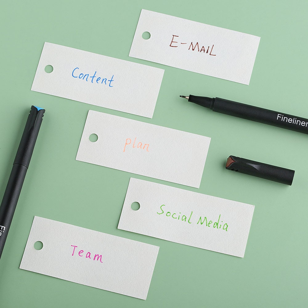 48 color fineliner pens set for journals and art, displayed with labeled tags reading "Content," "E-MAIL," "plan," "Social Media," and "Team," on a green background.