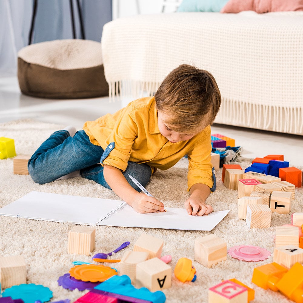 Child drawing with 72 Colors Dual Tip Brush Marker Set on white paper, surrounded by colorful building blocks and toys on beige carpet.