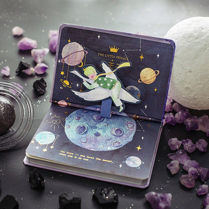 The Little Prince A5 3D Notebook with Bookmark featuring a pop-up illustration of the Little Prince flying among planets and stars, surrounded by gemstones and celestial decor.