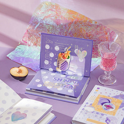 Emotional Palette A5 notebook with bookmark and 3D pages in purple theme, surrounded by decorative items including a half-cut plum, a crystal glass with pink drink, and colorful papers.