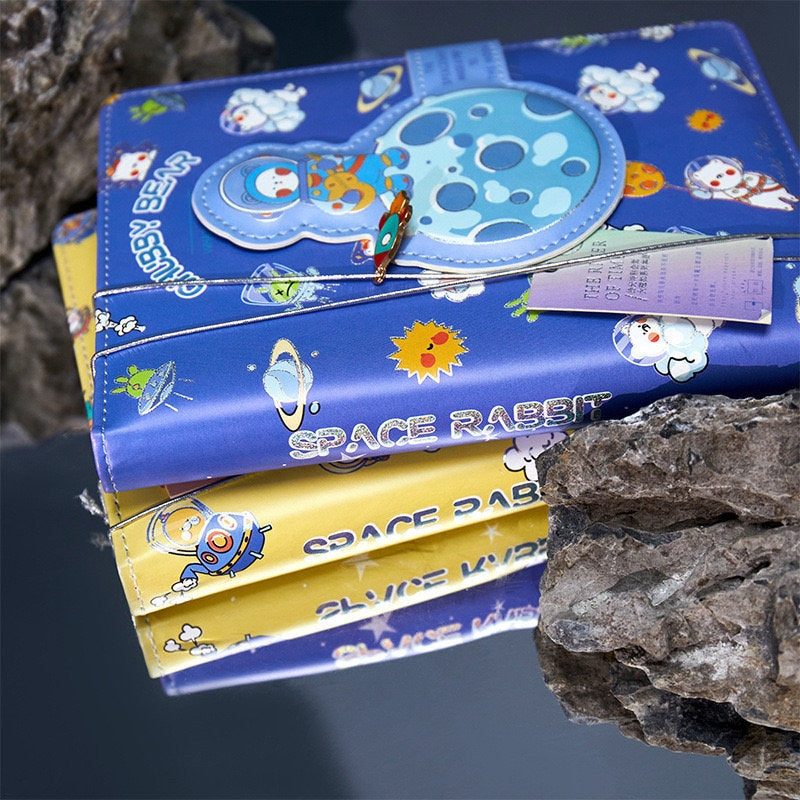 Spaceship Leather Journal with Bookmark featuring a Magnetic Buckle, blue and yellow 'Space Rabbit' design, nestled among rocks.