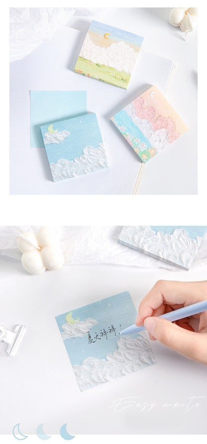 Flower Field Sticky Notes Set with 120 pieces, showcasing three sticky notes with colorful designs featuring floral fields, clouds, and crescent moons. A hand is writing on one of the sticky notes with a blue pen. Perfect for jotting down notes in a cute, kawaii style.