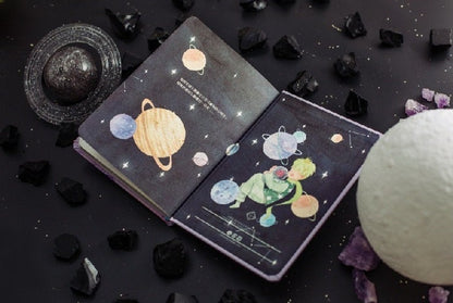 The Little Prince A5 3D Notebook with Bookmark featuring an open spread displaying colorful planets and The Little Prince illustration on a dark starry background, surrounded by black and purple decorative stones