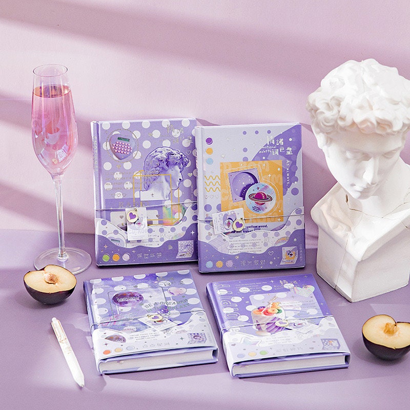 Emotional Palette A5 Notebook with Bookmark and 3D Pages in kawaii pastel purple design, surrounded by a glass of pink beverage, a white statue bust, a pen, and sliced fruit on a lavender surface.