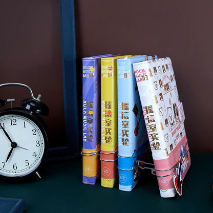 Rock Music Leather Journal with Magnetic Buckle, 224 Pages, standing vertically next to various colorful journals on a shelf beside a vintage black alarm clock.