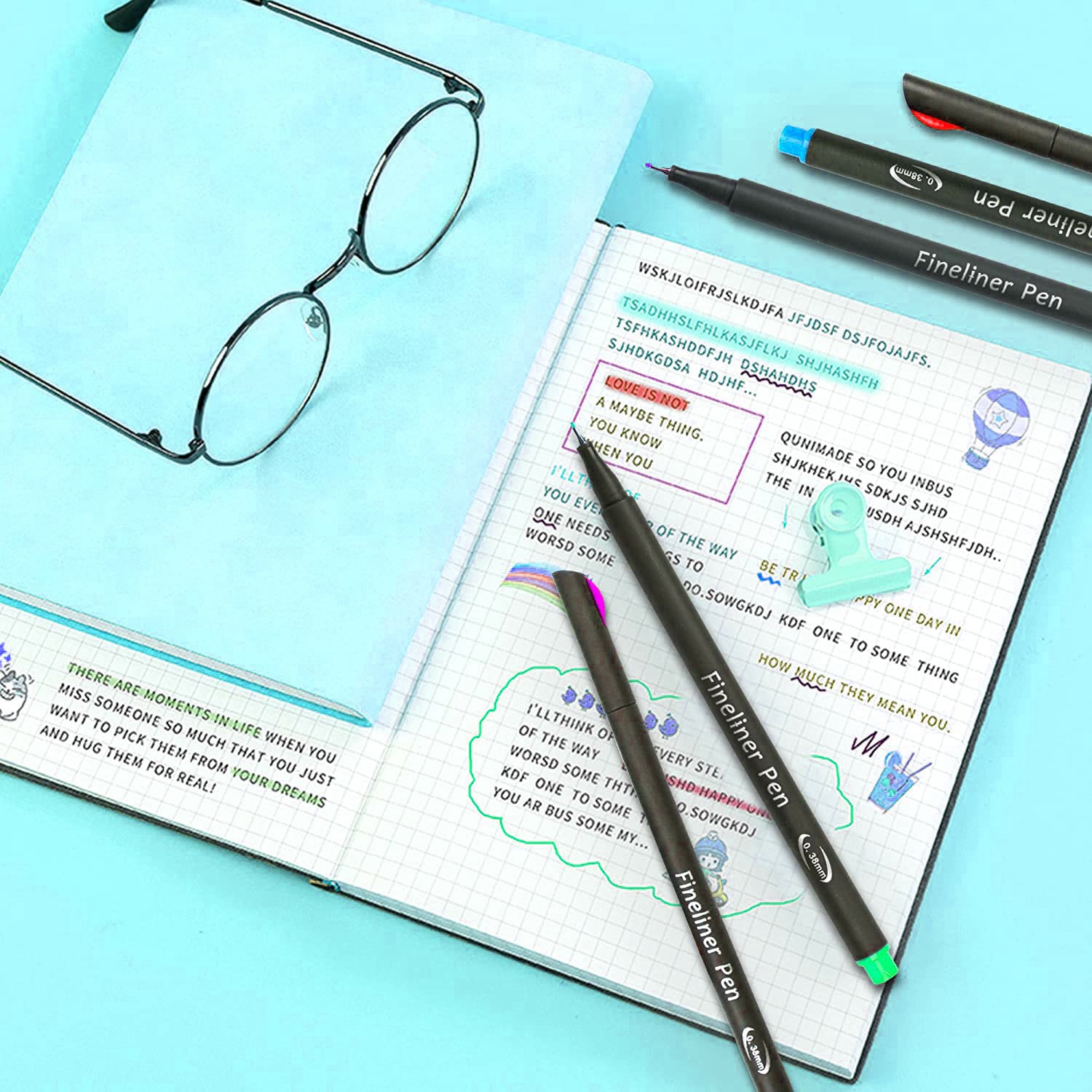 36-Color Fineliner Pens Set laid out with a journal featuring colorful drawings and text, black-rimmed glasses, and a teal binder clip on a light blue surface. Perfect for journaling, drawing, and note-taking.