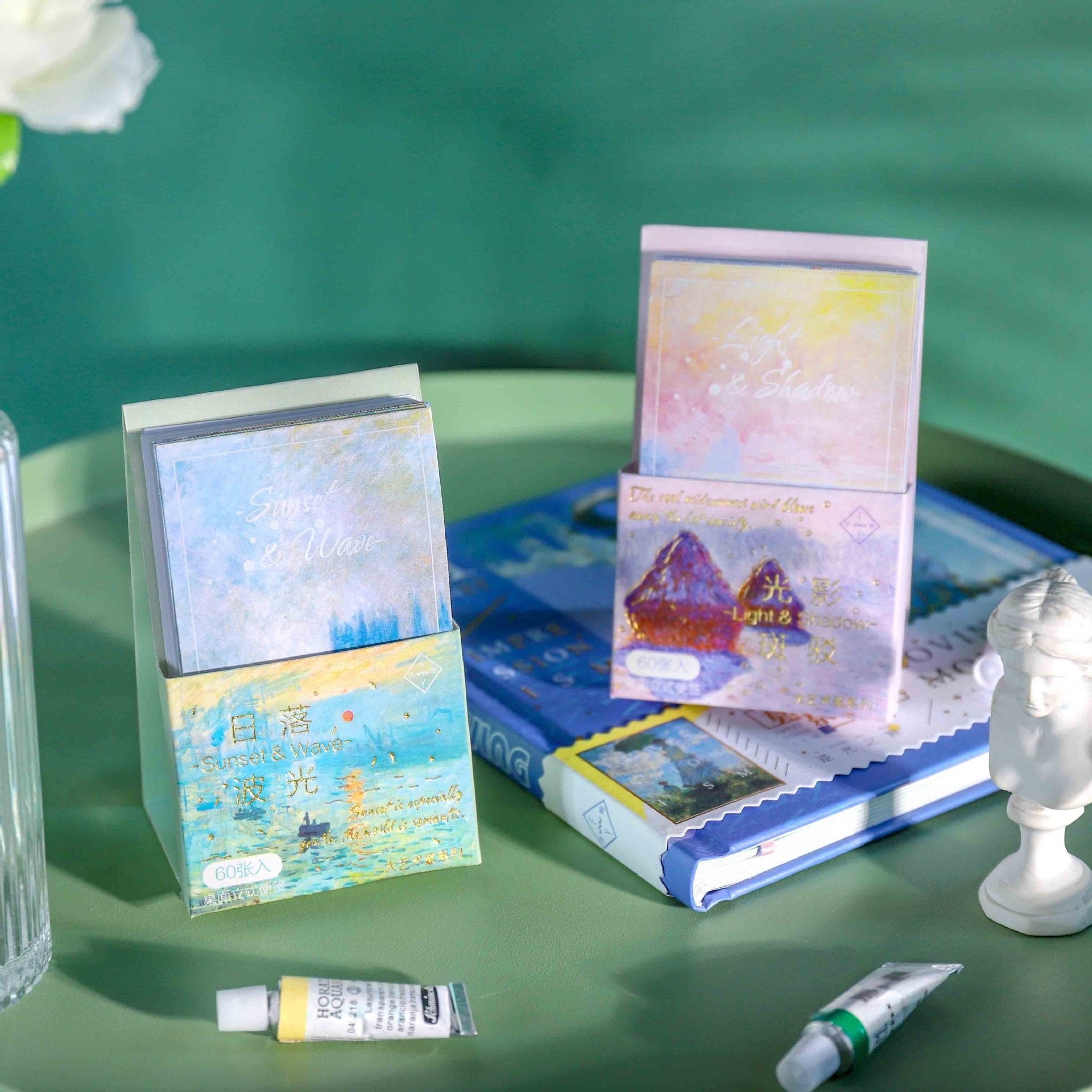 Sunset Wave Sticky Notepad with Stand - 60 Pages featuring a pastel ocean wave design, displayed alongside other stationery items and art supplies on a green surface.