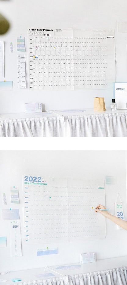 2022 Yearly Planner - Goal Tracker and Organizer displayed on a wall with months labeled from January to December, featuring goal-setting spaces, surrounded by a notebook, sticky notes, and other stationery items for effective planning and organization.