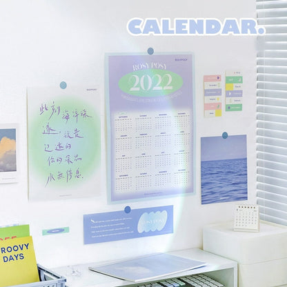 2022 Goal Tracker Calendar with Stickers in pastel colors, displayed on a white wall along with other decorative notes and photos, next to a window with blinds.