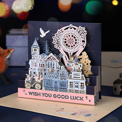 Starlight Studio Pop Up 3D Greeting Card featuring an intricate design of a whimsical village with a Ferris wheel and a "Wish You Good Luck" message in delicate pastels against a dark background.