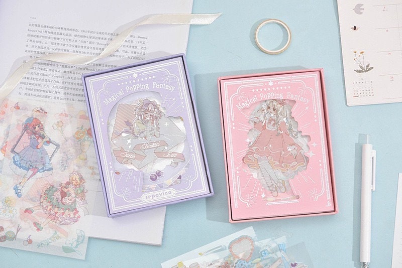 Magical Silver Foil Stickers for Planners and Crafts featuring two boxes in kawaii style packaging, one in purple and one in pink, surrounded by stationery items like decorative tape, pen, and planning sheets