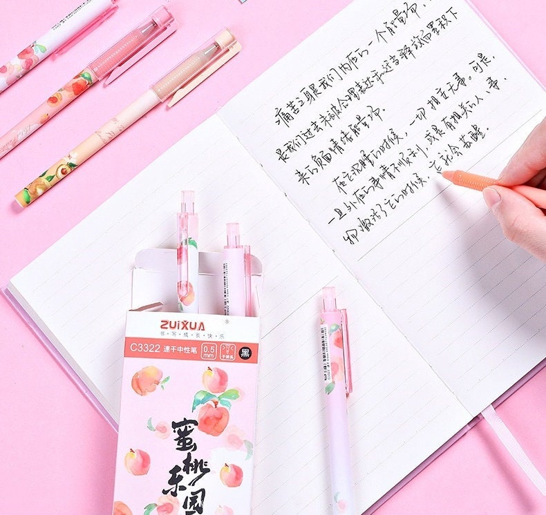 Peach Scented 0.5mm Kawaii Gel Pen set with pink and peach-themed designs, displayed on a pink background with an open notebook showing handwritten text and a hand holding one of the pens.