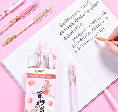 Peach Scented 0.5mm Kawaii Gel Pen set with pink and peach-themed designs, displayed on a pink background with an open notebook showing handwritten text and a hand holding one of the pens.