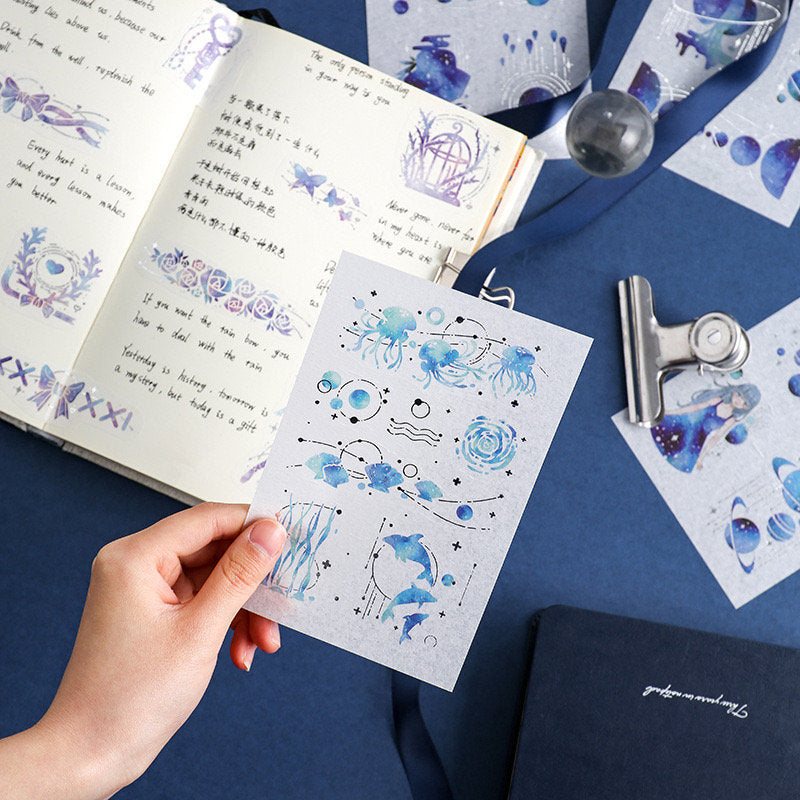 Dream Star Silver Foil Washi Sticker Set featuring celestial and ocean-themed designs in blue and silver, showcased with a hand holding one sheet, surrounded by a journal, additional sticker sheets, and stationery items.