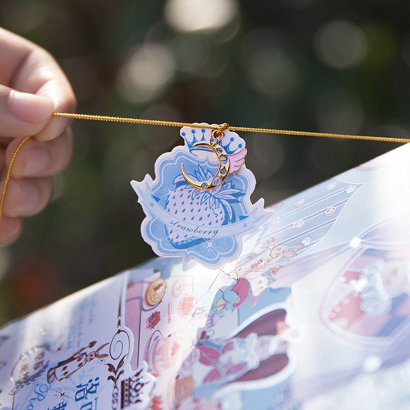 Close-up of Rococo Waltz Leather Journal with Metal Bookmark, featuring a delicate strawberry-themed charm and intricate illustrations, perfect for kawaii stationery lovers.