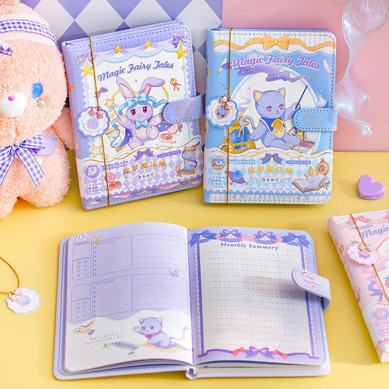 Magic Fairy Tales Journal in pink and blue leather, A5 size, 196 pages, featuring cute animal illustrations, open to monthly summary pages, placed on a pastel-themed desk with a plush toy.