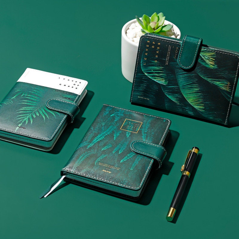 Tropical Leaf Journal with magnetic buckle, green leather cover featuring leaf designs, displayed alongside a black and gold pen and a small potted plant.