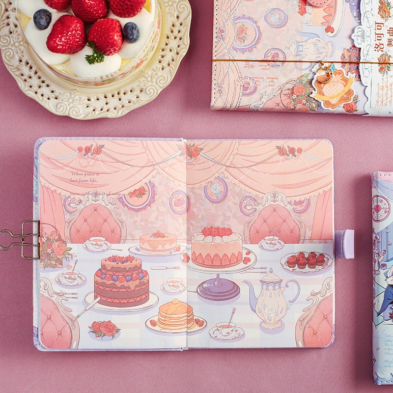 Rococo Waltz Leather Journal with Metal Bookmark featuring an elegant cake and tea party illustration, displayed on a pink cloth background next to a dessert plate and matching stationery items.