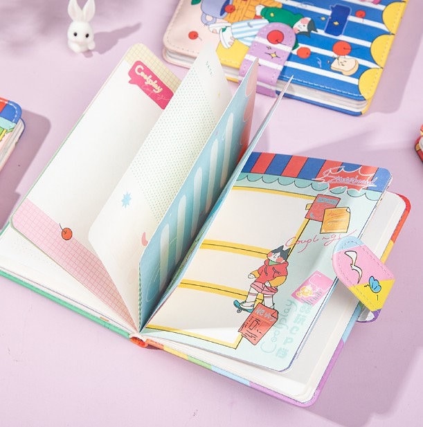 Coolplay A6 Magnetic Leather Journal in blue, open to show colorful, illustrated pages and decorative inserts on a pastel pink surface with other kawaii stationery items in the background.