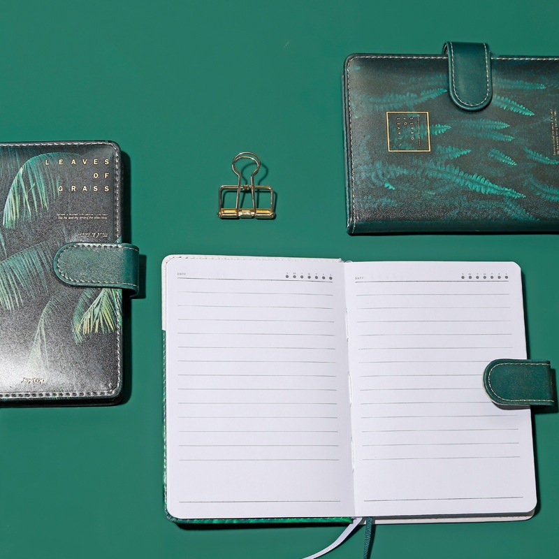 Tropical Leaf Journal with Magnetic Buckle, featuring a green leather cover and open lined pages, displayed with a gold binder clip on a matching green background.