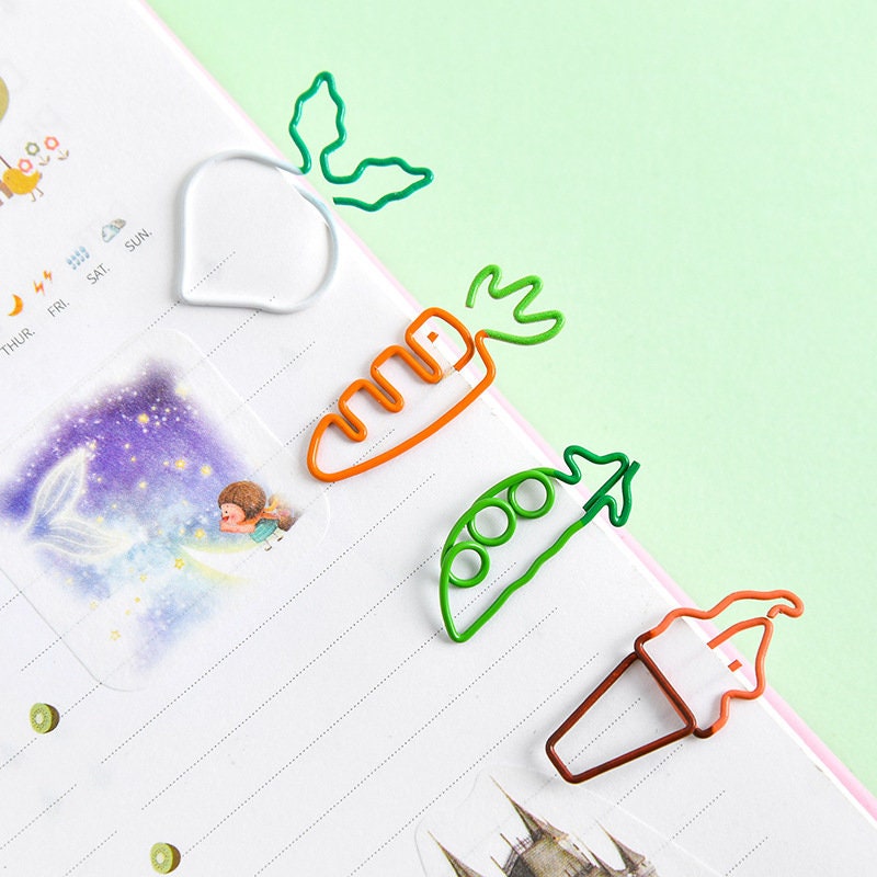 Creative vegetable clips in shapes of a radish, carrot, pea pod, and ice cream cone, attached to a notebook. Cute stationery and office supplies in kawaii style.