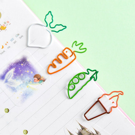 Creative vegetable clips in shapes of a radish, carrot, pea pod, and ice cream cone, attached to a notebook. Cute stationery and office supplies in kawaii style.