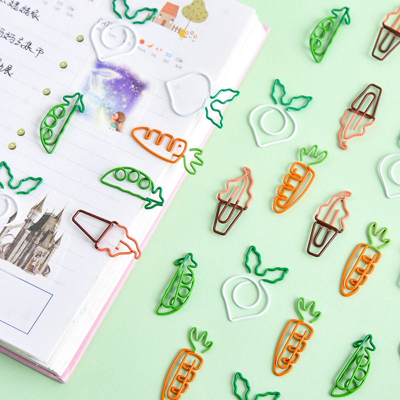 Creative vegetable-shaped paper clips in carrot, pea, radish, and other designs placed on an open planner with illustrations. Cute stationery and office supplies.